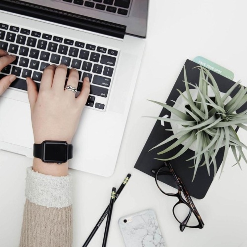 overhead shot of woman working on a laptop at a round table with pencils, her cell phone, glasses, notebook and a potted plant