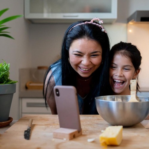 mother and young daughter smile at their phone as they cook in the kitchen